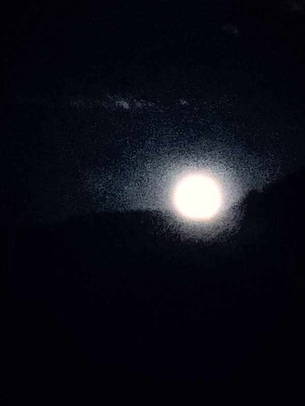 A bright, round moon set high against a black mountain ridge and a dark sky.