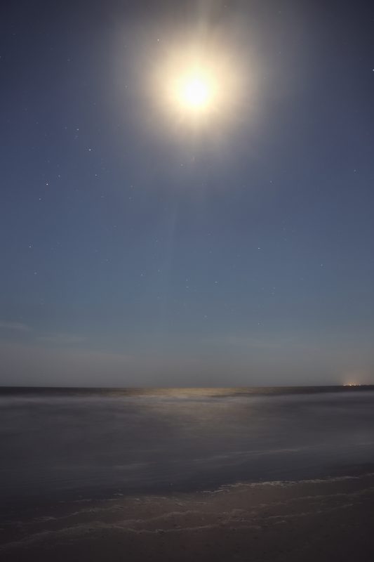 A bright moon high in a pale blue sky over a hazy beach and calm surf at dusk.