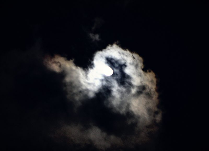 A moon glowing behind a bright, billowing cloud in a black sky, the cloud edges lit white and blue.