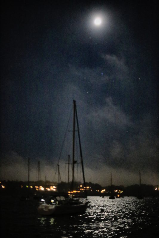 A bright moon high over a harbour at night, a sailboat mast reaching up toward it and lights shimmering on the water.