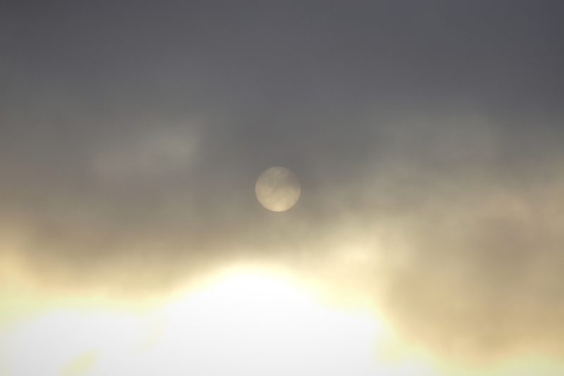 A dim moon hidden in a pale, glowing haze of cloud above a bright, misty horizon.