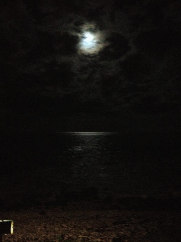 A moon glowing through dark clouds above a black sea, a faint path of light on the water below.