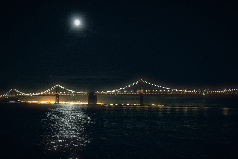 A bright moon above the dark bay, the Bay Bridge strung with glowing lights and their reflections across the water.