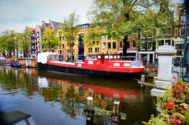 A red houseboat on a canal reflecting gabled houses, red flowers in the corner.