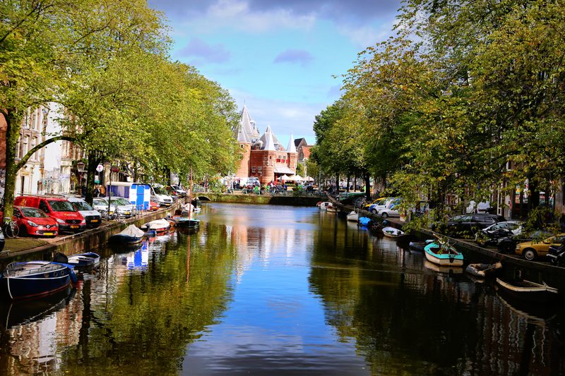 Trees and canal houses reflected in a still canal under blue sky.