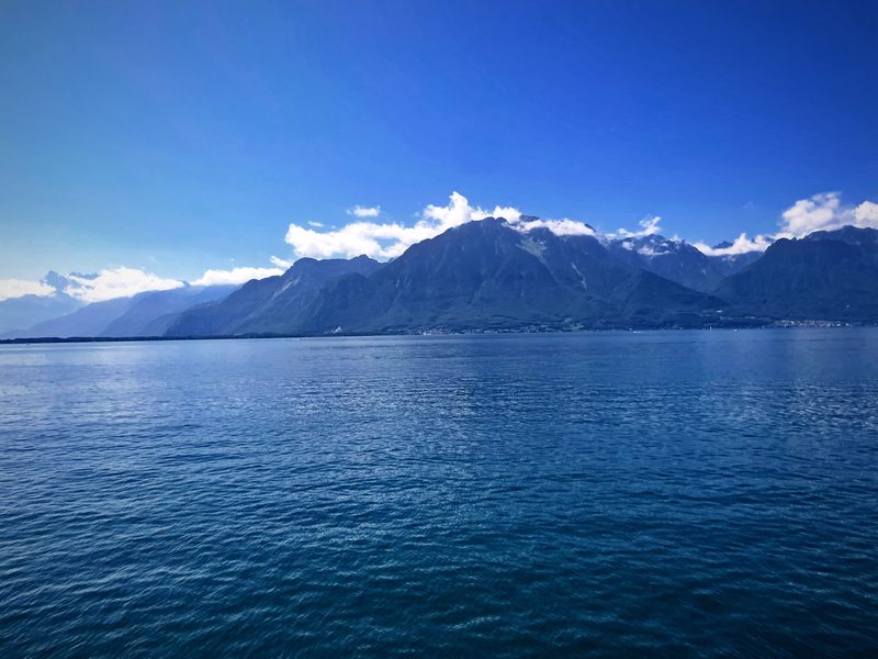 Deep teal Lake Geneva water stretching to snow-streaked peaks under bright clouds.