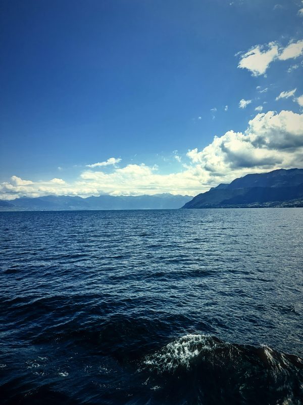 Dark blue lake water rolling toward hazy mountains under white clouds.