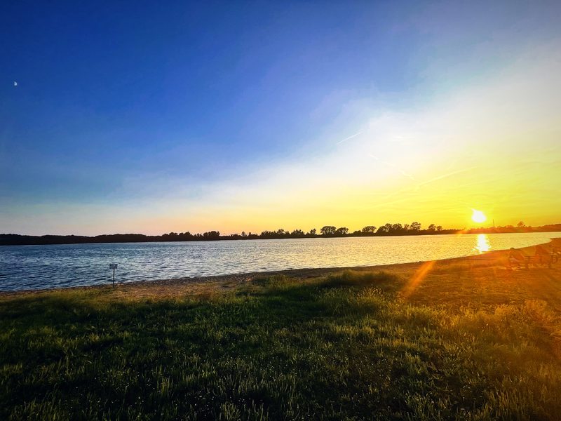 A golden sun setting over a still lake beside a curve of green grass.