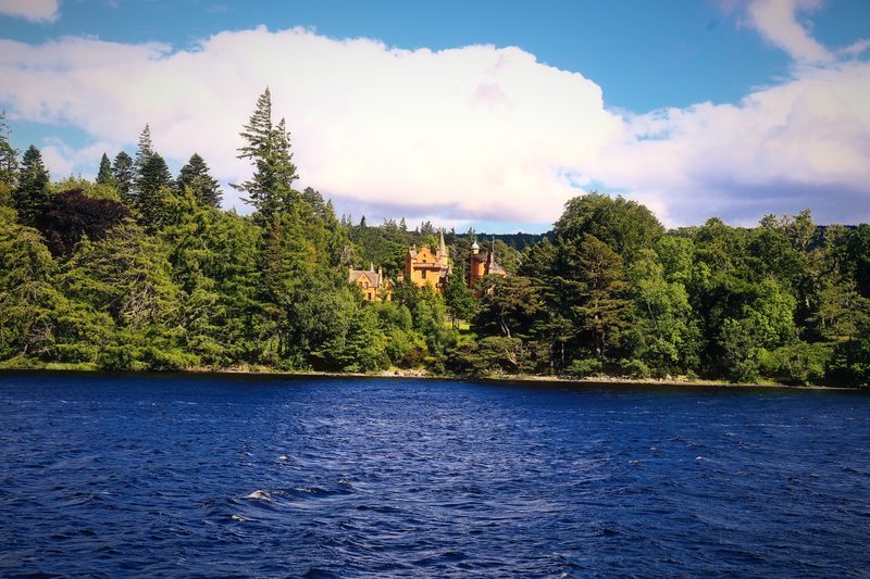 Deep blue loch water below a wooded shore and a pink house under big white clouds.