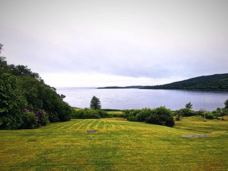 A striped green lawn rolling down to a pale silver bay and low hills.