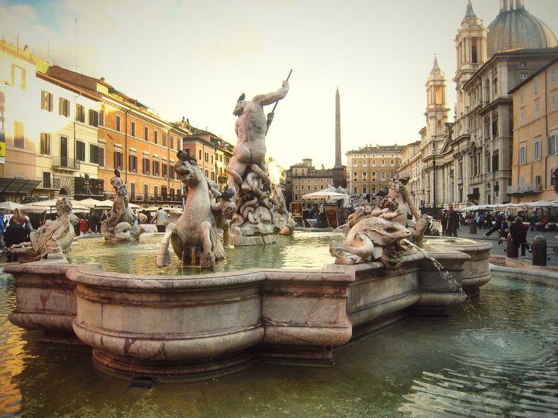 A baroque fountain with sculpted figures in Piazza Navona, buildings and a dome behind.