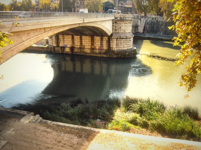 A bridge over the Tiber with sunlight on the water and the bank below.