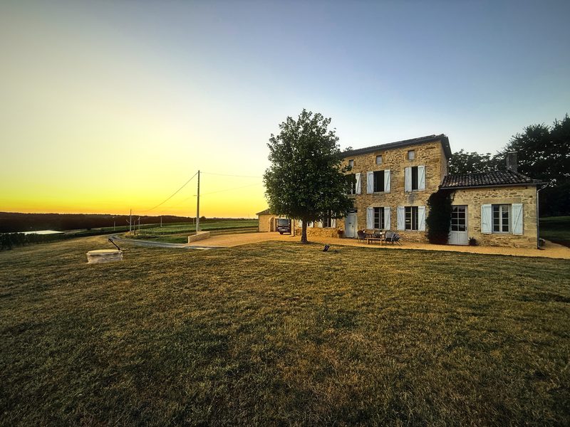 A stone farmhouse and a single tree on a dry lawn as the sun sets gold on the horizon.