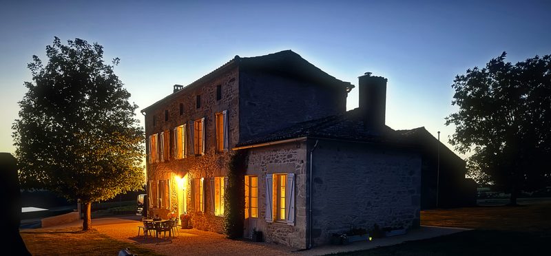 A stone house glowing with a lit lantern at dusk, framed by trees under a deepening sky.