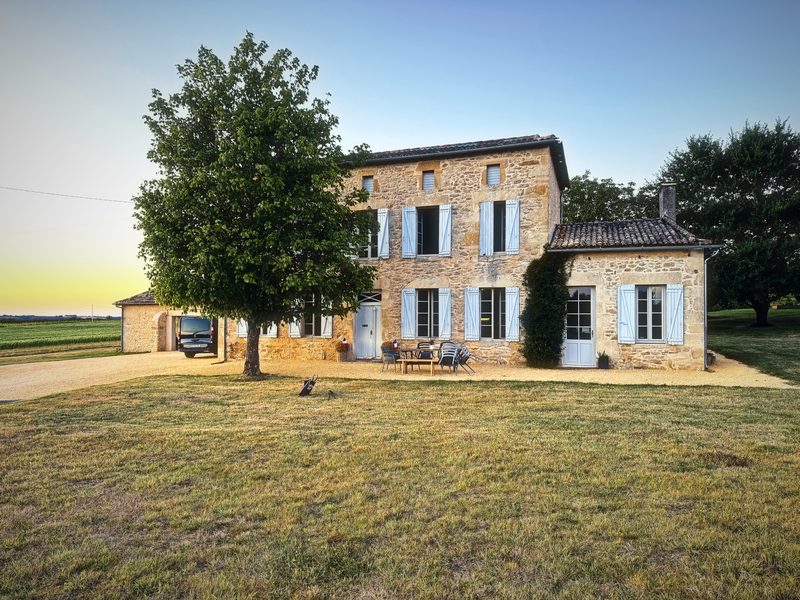 A stone farmhouse with blue shutters and a big tree, lit warm by the low evening sun.