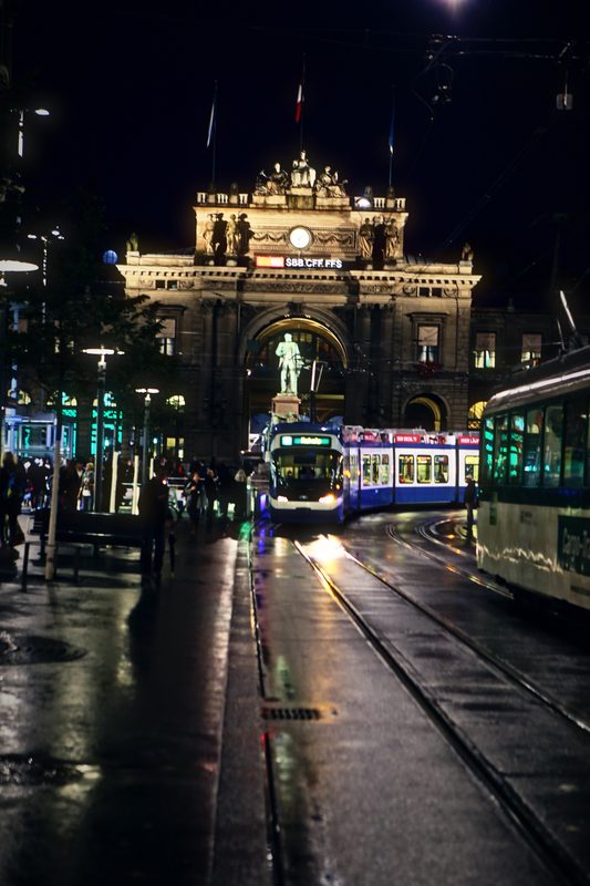 Floodlit stone station façade at night with a statue and wet tram tracks in front.