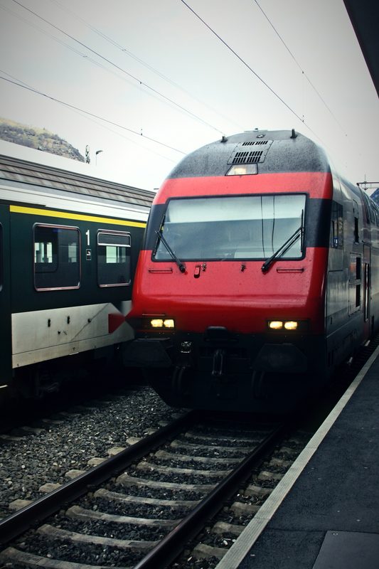 Red Swiss electric locomotive arriving at a platform beside a waiting train.