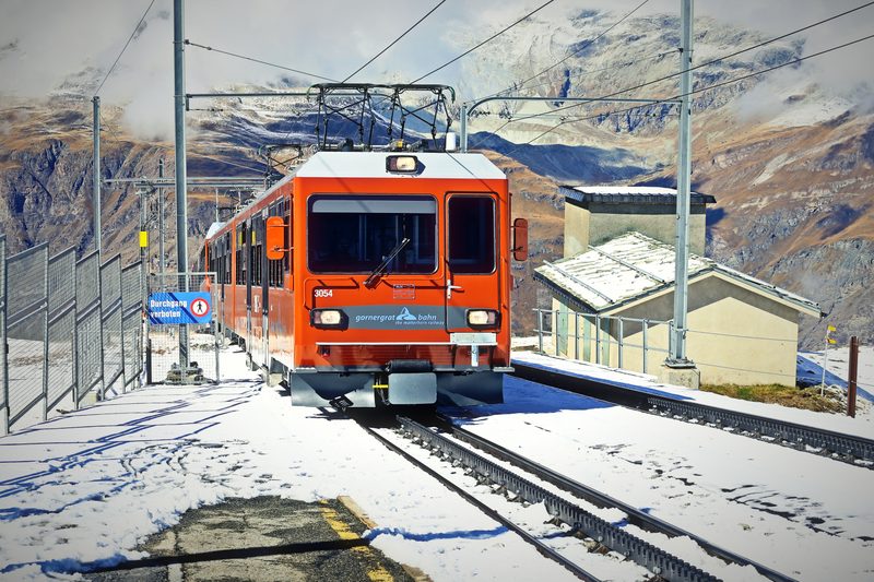Orange mountain train at a snowy station with peaks behind.