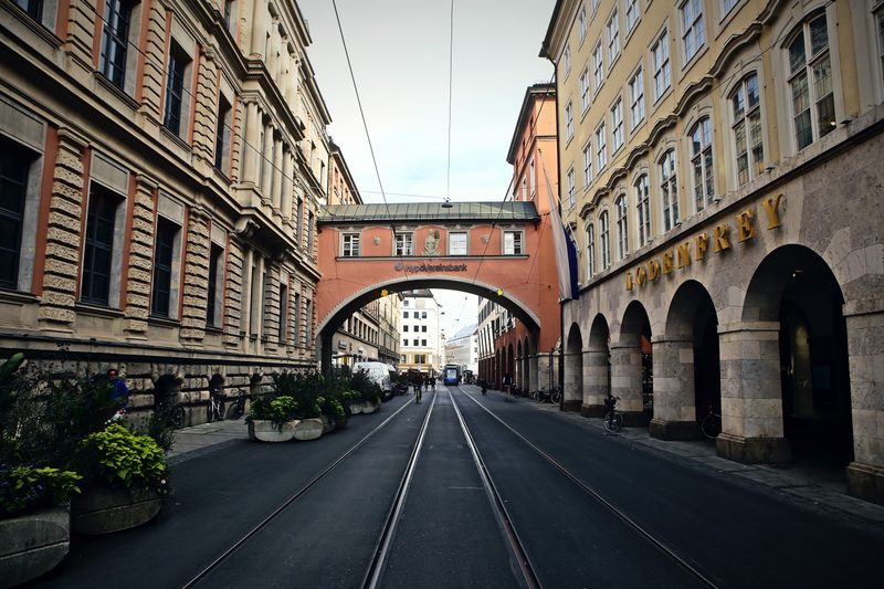 Street with tram rails running under a covered bridge between old buildings.