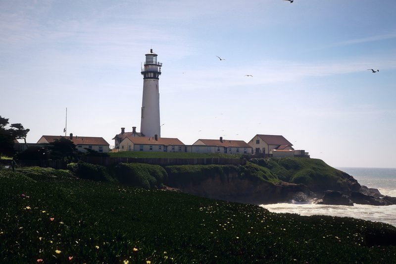 Tall white lighthouse above red-roofed station buildings and surf on the Pacific coast.