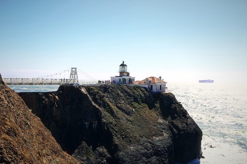 Small lighthouse at the end of a dark cliff reached by a footbridge, with a ship out at sea.