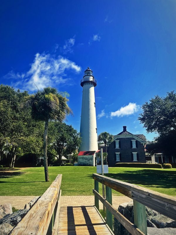 Wooden boardwalk leading to a white lighthouse and keeper's house under summer clouds.