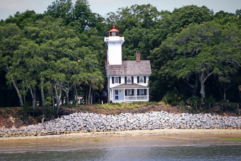 Small lighthouse on a white keeper's house above a stone seawall and live oaks.