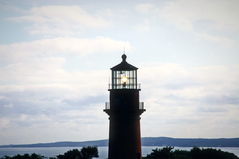 Dark lighthouse tower with its lamp glowing, above trees and open water.