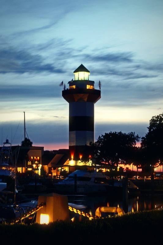 Striped lighthouse with its lamp on above a marina as the last sunset color fades.
