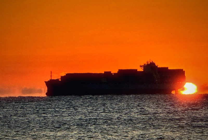 A container ship in silhouette crossing a burning orange sea as the sun sets at the waterline.