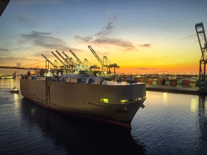 A huge car-carrier ship easing through a harbor channel beneath container cranes and a gold sunset.