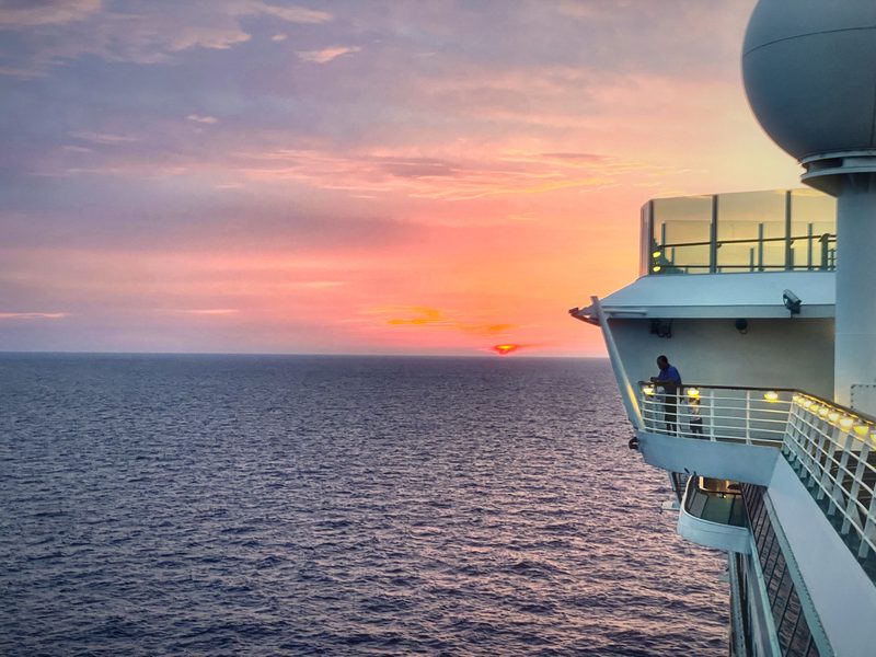 The side of a cruise ship at sunset, a lone figure at the rail and a pastel sky over open water.