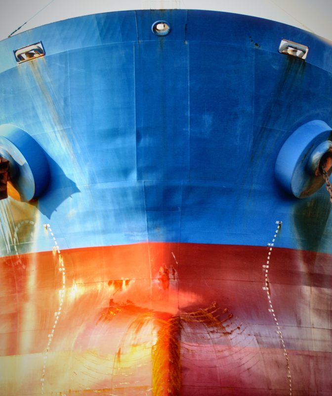 The bow of a ship painted bright blue over red, with anchors in their hawse pipes and draft marks running down the hull.