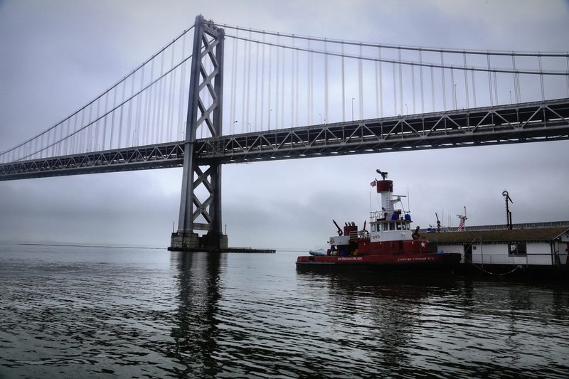 A red fireboat moored beneath a long suspension bridge on still grey water.