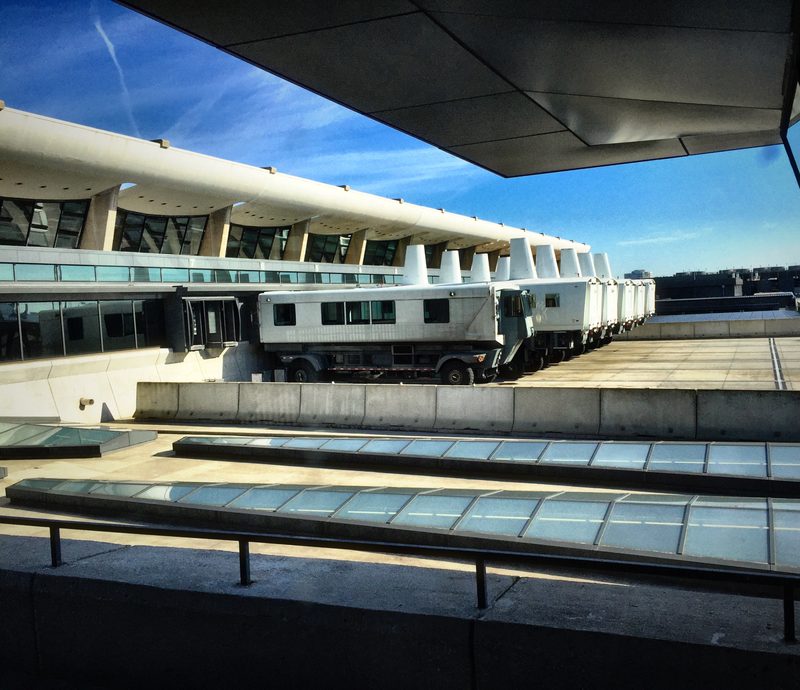 A line of white airport mobile lounges parked beneath a sweeping concrete terminal roof.
