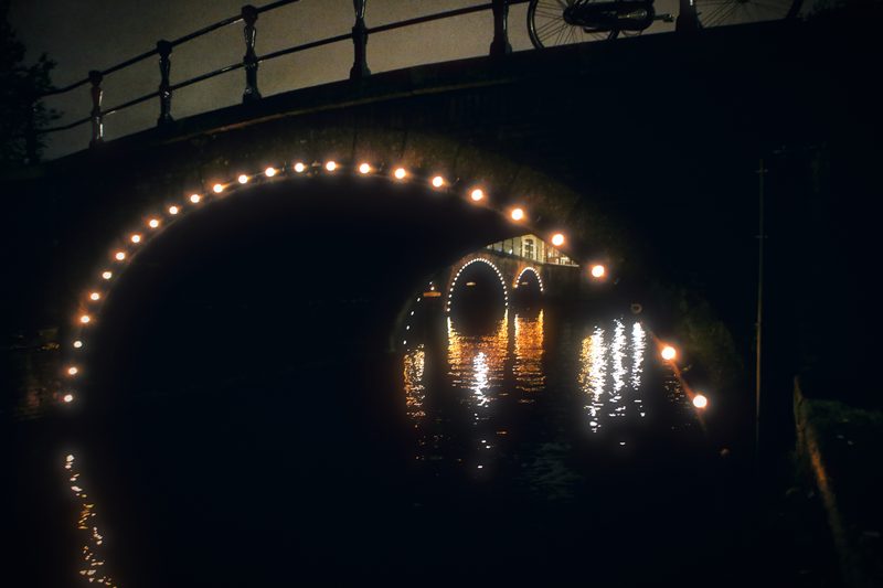 A canal bridge outlined in light bulbs reflected as a ring in black water.