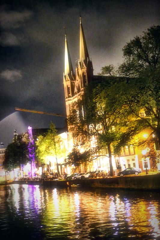 A Gothic church with twin spires lit at night above a canal reflecting violet and gold.