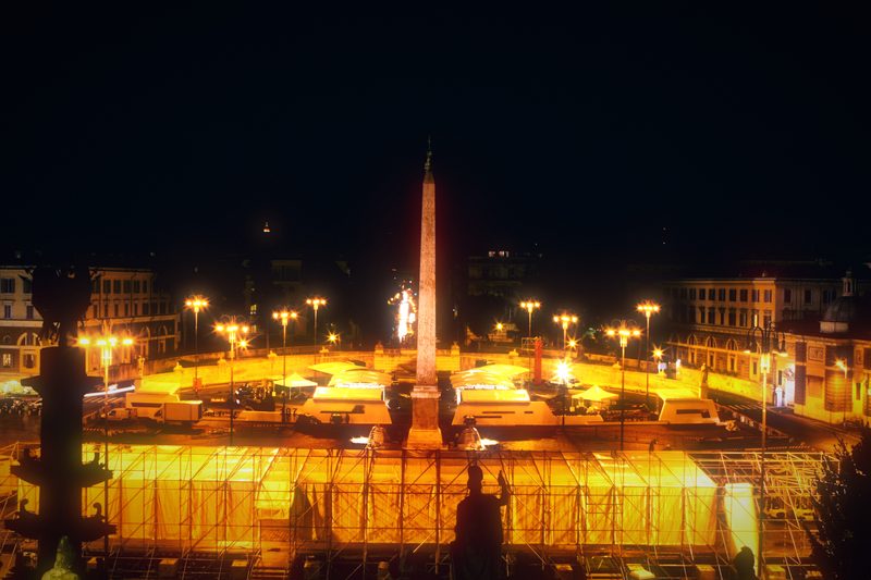 Piazza del Popolo at night with its obelisk and a sweep of lights.