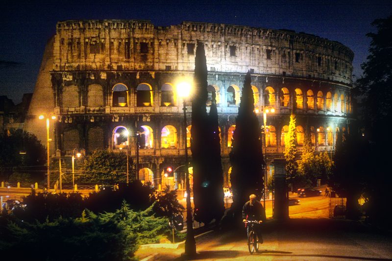 The Colosseum at night behind a dark cypress tree.