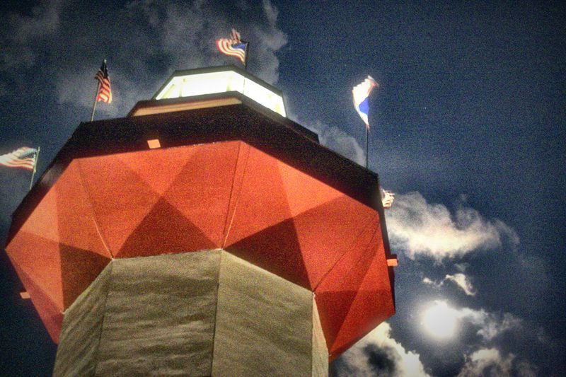 A faceted red-paneled tower crowned with flags under a dark, stormy sky.