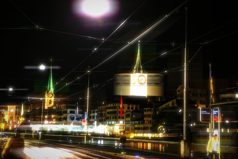 Night tram stop with glowing clock, church spire and blurred lights.