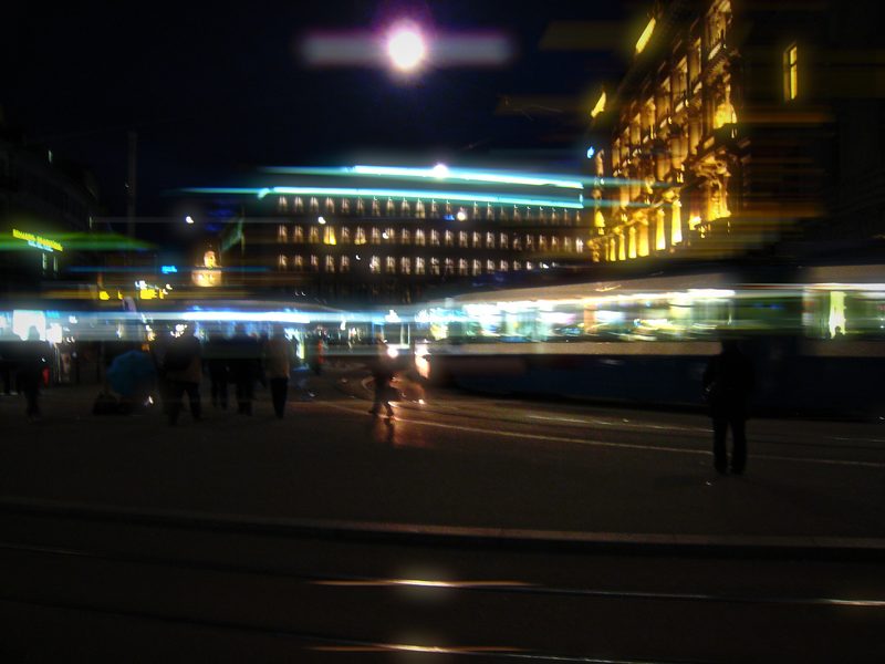 Night square with trams streaking past in cyan light and lit buildings.