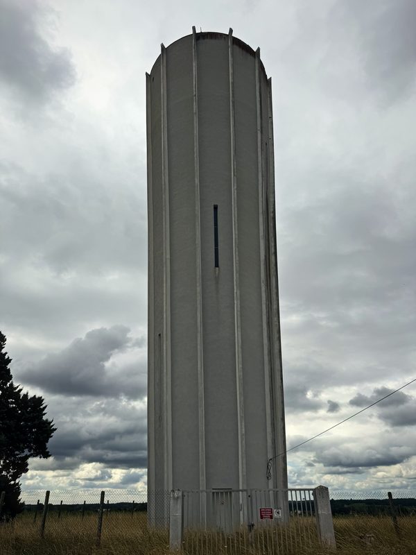 A tall concrete village water tower rising from the fields against a grey, broken sky.