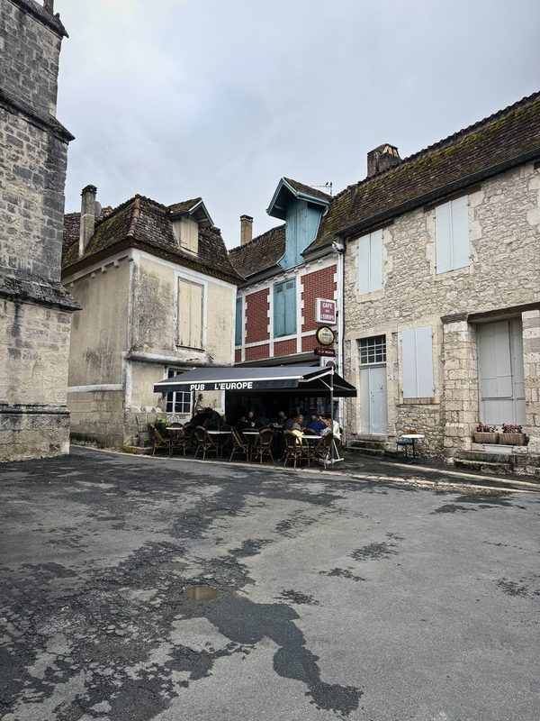 A bend of an old stone village street with shuttered houses and a café awning.