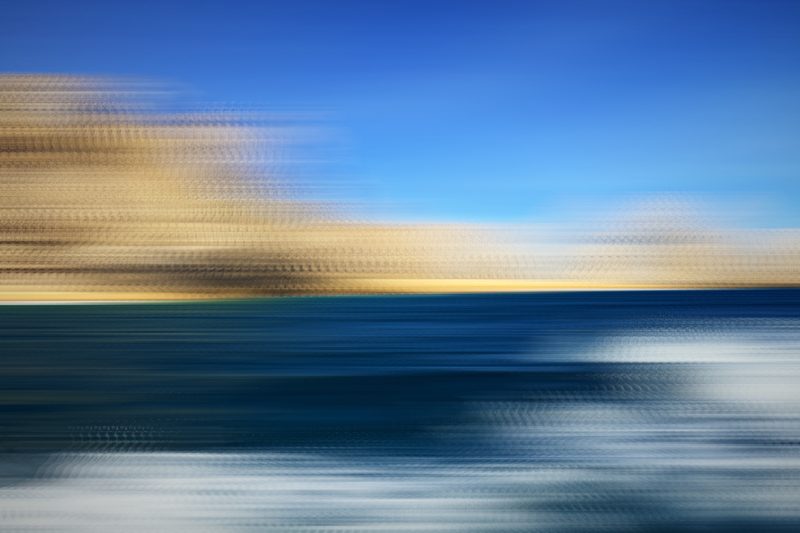 Horizontal streaks of tan cliffs, blue sea and white surf.