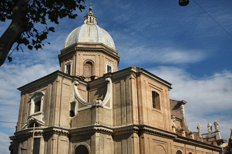 A domed church in warm stone against blue sky, trees at its edge.