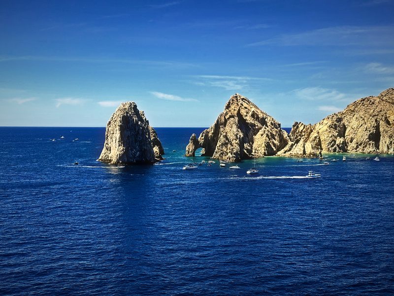 A rock arch and lone pillar rising from deep sapphire water under a blue sky.
