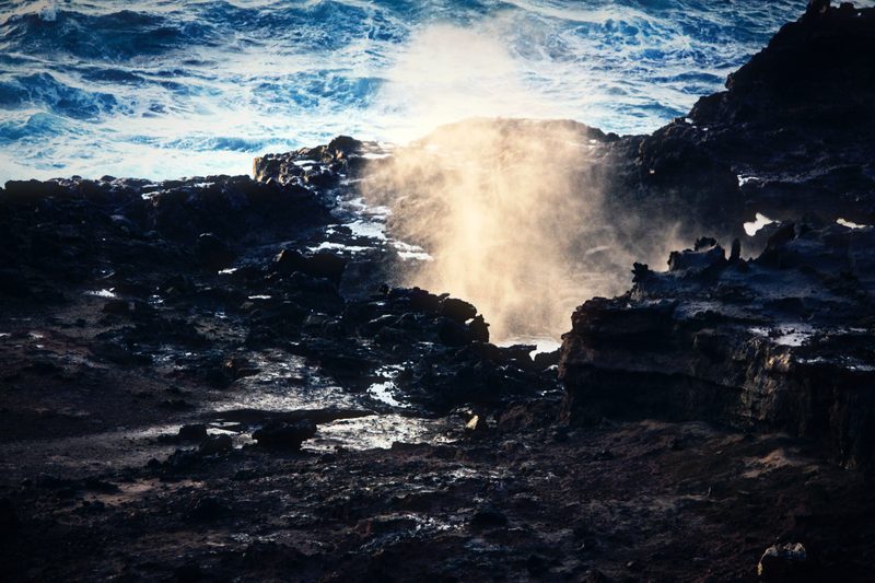 A plume of sea spray bursting up through black volcanic rock.