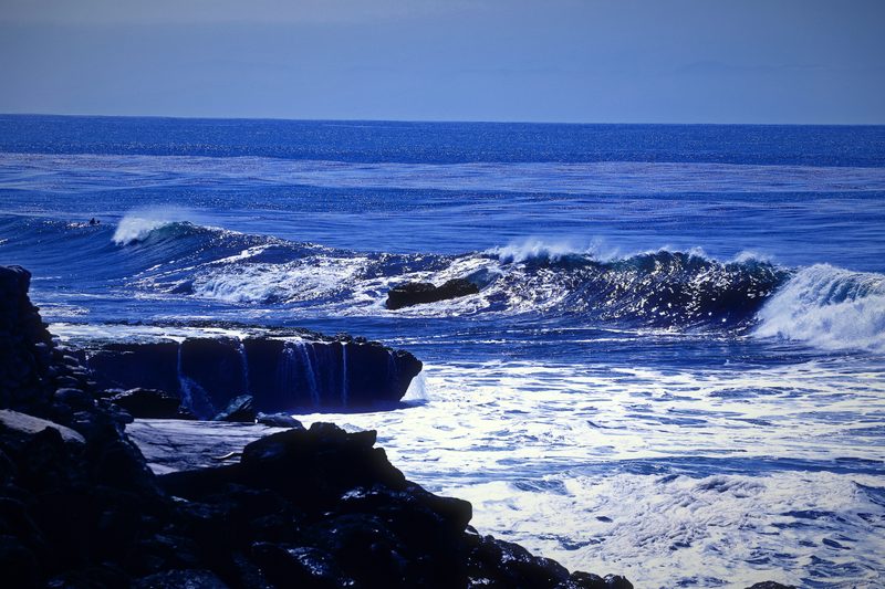 A flat dark rock taking a hit of white surf on a blue day.