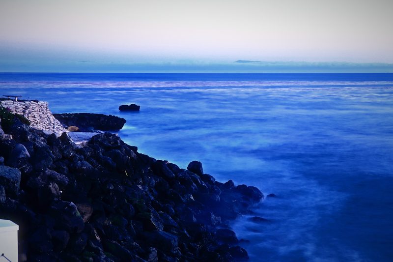 Dark rocks running into a smooth blue sea under a pale sky.